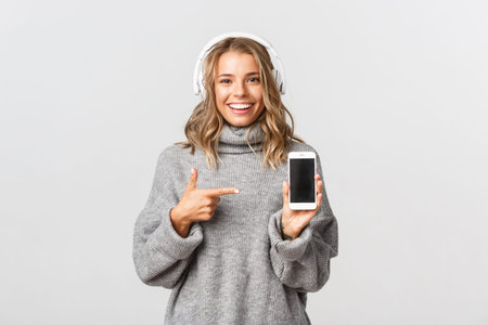 Studio shot of attractive blonde girl in grey sweater, listening music in wireless headphones,...