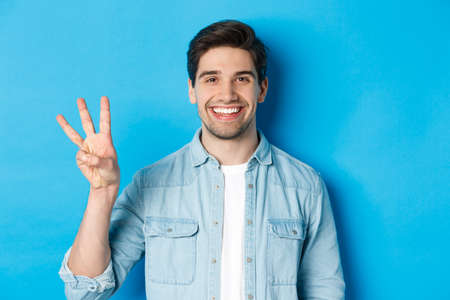 Close-up of handsome man smiling, showing fingers number three, standing over blue backgroundの写真素材