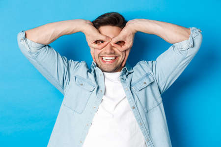 Young caucasian guy showing funny expression, making superhero mask with fingers on eyes, smiling happy, standing over blue backgroundの写真素材