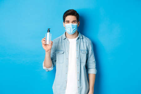 Concept of covid-19, pandemic and social distancing. Young man in medical mask showing hand sanitizer, standing against blue backgroundの写真素材