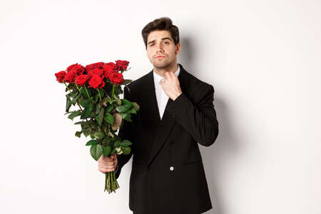 Image of elegant and sassy man in black suit, looking confident and holding bouquet of red roses, going on a romantic date, standing against white backgroundの写真素材
