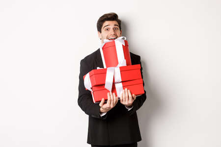 Concept of christmas holidays, celebration and lifestyle. Image of happy man in suit carry presents for new year, holding boxes with gifts and smiling, standing against white backgroundの写真素材