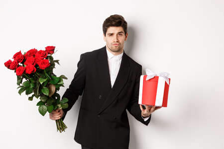 Concept of holidays, relationship and celebration. Handsome and confident man in black suit, going on a date, holding bouquet of roses and present, standing against white backgroundの写真素材