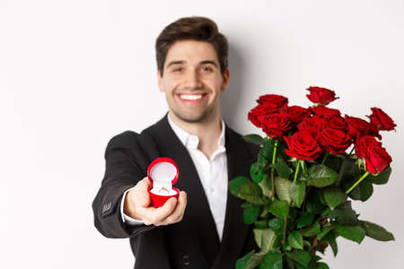 Close-up of attractive man in suit, holding bouquet of roses giving an engagement ring, proposing to girlfriend, standing against white backgroundの写真素材