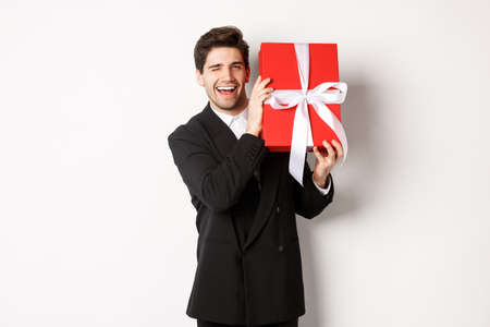 Concept of christmas holidays, celebration and lifestyle. Attractive man in black suit, holding new year gift and smiling, standing with a present over white backgroundの写真素材