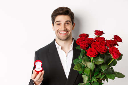 Close-up of attractive man in suit, holding bouquet of roses and engagement ring, making proposal, standing against white backgroundの写真素材