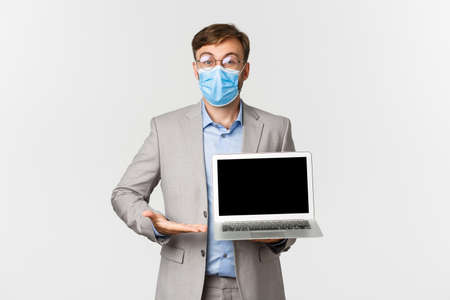 Concept of work, covid-19 and social distancing. Portrait of impressed boss in medical mask, showing something amazing on laptop screen, demonstrating presentation, white backgroundの写真素材
