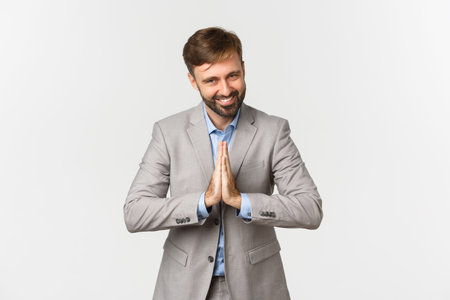 Portrait of happy bearded businessman in grey suit, thanking for something, holding hands pressed together and bowing with grateful expression, standing over white backgroundの写真素材