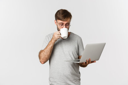 Image of male freelancer working with laptop, drinking coffee, standing in gray t-shirt and glasses...