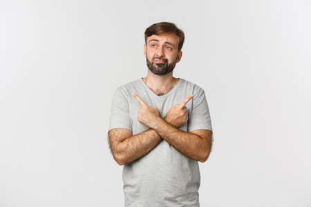Portrait of indecisive beaded man in gray t-shirt, making decision, pointing fingers sideways at two...