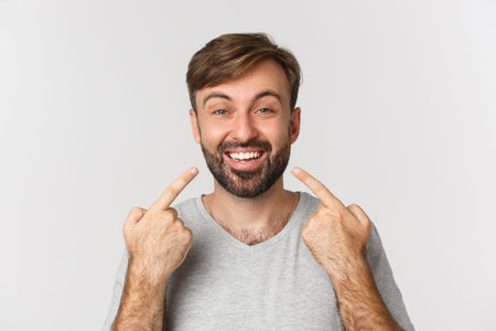 Close-up of handsome smiling man recommending dental clinic, pointing at his perfect white teeth,...