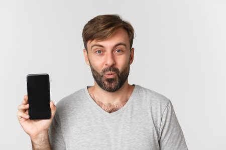 Close-up of amazed handsome man in gray t-shirt, showing mobile phone screen, recommending app or...