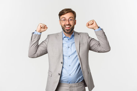 Portrait of happy businessman with beard, wearing grey suit and glasses, raising hands up in hooray...
