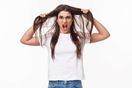 Waist-up portrait of bothered and annoyed, angry pissed-off young girlfriend losing temper, pulling hair from head and screaming being furious and outraged, white backgroundの写真素材