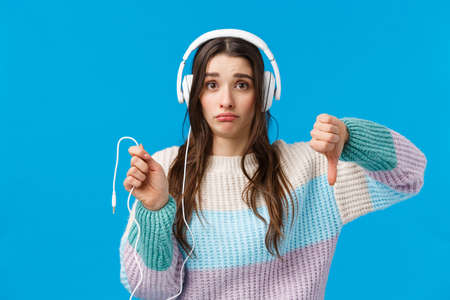 Sad and displeased, gloomy cute brunette woman in big headphones, grimacing disappointed, showing thumbs-down and holding dropped wire of earphones, standing blue backgroundの写真素材