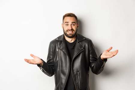 Confused bearded man in black leather jacket asking for advice, looking clueless and shrugging,...