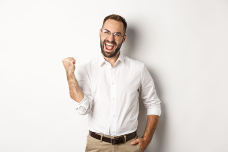 Successful businessman rejoicing, making fist pump and saying yes, achieve goal, standing over white...