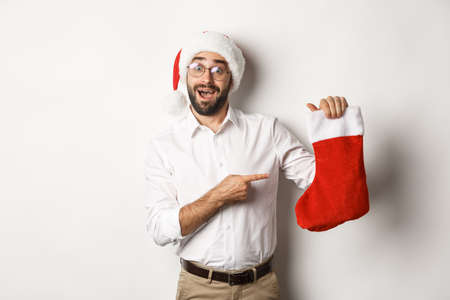 Merry christmas, holidays concept. Happy adult man receive gifts in xmas sock, looking excited, wearing santa hat, white backgroundの写真素材