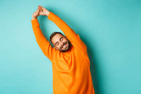 Image of handsome young man stretching hands and smiling after good rest, standing in orange sweater over light blue backgroundの写真素材