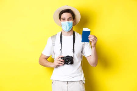 Concept of covid-19, travelling and quarantine. Happy man tourist with camera, showing passport and tickets for vacation, going on trip during pandemic, yellow backgroundの写真素材