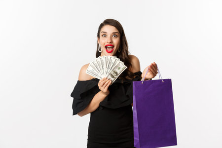 Image of excited beautiful woman in black dress going shopping, holding bag and dollars, standing...
