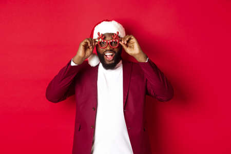 Funny Black man celebrating New Year, wearing party glasses and santa hat, having fun over red backgroundの写真素材