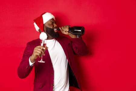 Christmas, party and holidays concept. Image of young Black man in santa hat drinking champagne from bottle, getting drunk on New Year celebration, standing over red backgroundの写真素材