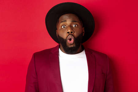 Close-up of surprised Black man staring at camera with wonder, standing against red backgroundの写真素材