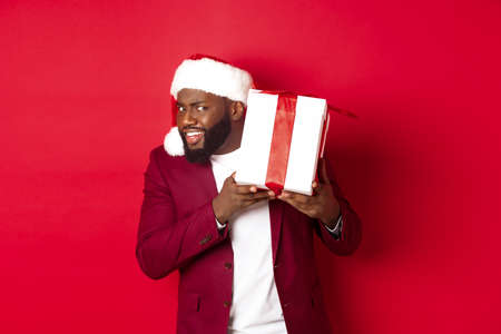 Christmas. Curious Black man in santa hat shaking new year gift, wonder what inside box, standing against red backgroundの写真素材