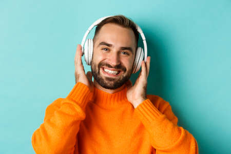 Close-up of handsome modern man listening music in headphones, standing in orange sweater over turquoise backgroundの写真素材