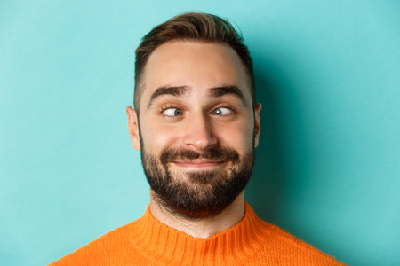 Headshot of funny caucasian man making faces, squinting and smiling silly, standing over light blue...