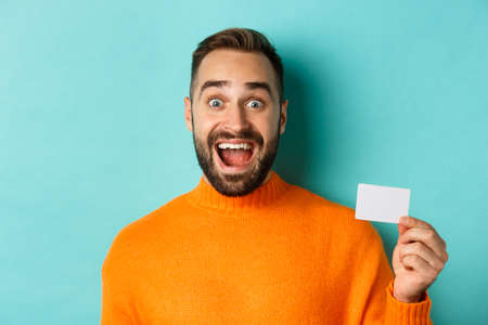 Close-up of excited caucasian man showing his credit card, smiling and staring amazed, standing in orange sweater against turquoise backgroundの写真素材
