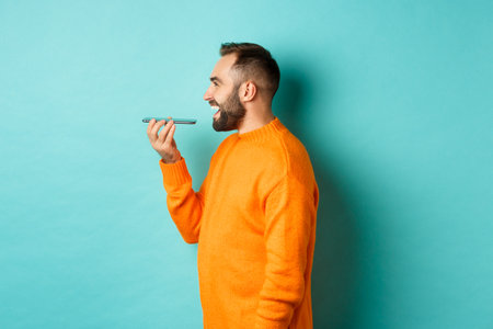 Profile of handsome bearded man recording voice message, holding mobile phone and talking on...
