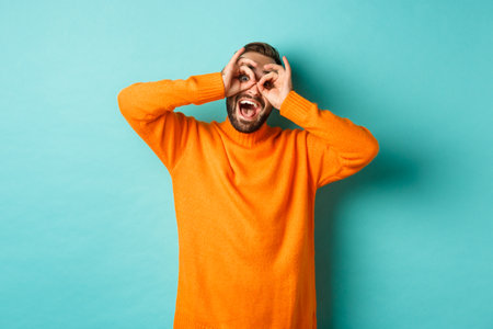 Image of funny and happy guy holding hands on eyes, looking through fake glasses, smiling amused, standing in orange sweater against turquoise backgroundの写真素材