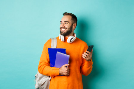 Education. Handsome male student with headphones and backpack, using mobile phone and holding notebooks, smiling happy, standing over turquoise backgroundの写真素材