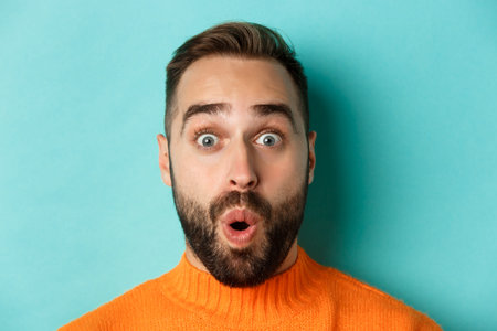 Headshot of handsome caucasian man with beard standing in orange sweater against turquoise...