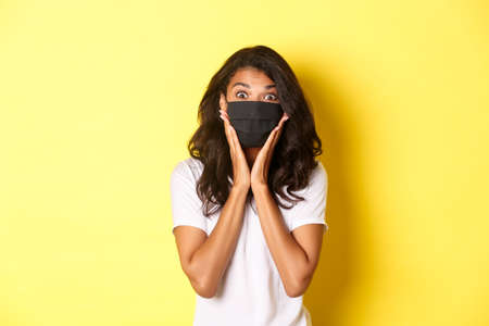 Concept of coronavirus, pandemic and lifestyle. Portrait of excited african-american woman in face mask, looking amazed at something cool, standing over yellow backgroundの写真素材
