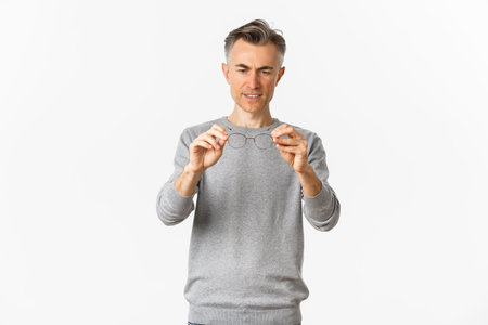 Portrait of middle-aged man in grey sweater, looking at glasses, standing over white background