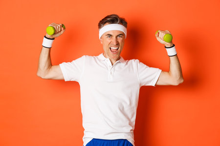 Portrait of confident middle-aged fitness guy, doing sports over orange background, flex biceps and...