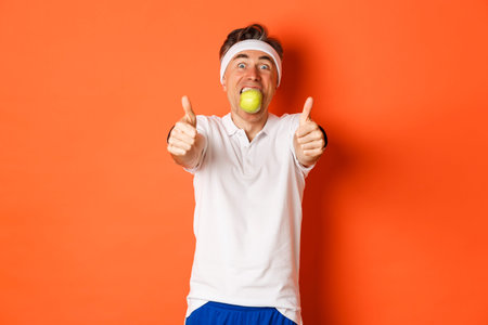 Image of funny middle-aged guy in fitness clothing, holding apple in mouth, showing thumbs-up,...