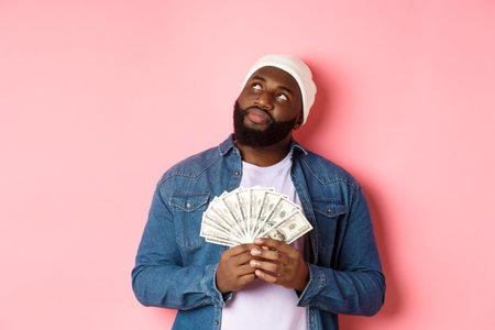 Dreamy african-american man thinking about shopping, holding money dollars and looking upper left...