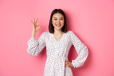 Pretty young asian woman in dress showing okay sign, praising and showing approval, looking satisfied, standing against pink backgroundの販売画像