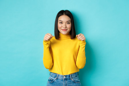 Beauty and fashion concept. Beautiful and stylish asian woman in yellow pullover, holding hands raised near chest as if holding banner or logo, standing over blue backgroundの写真素材