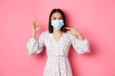 Coronavirus, social distancing and lifestyle concept. Cute asian woman pointing at face mask, showing okay sign, support using preventive measures from covid-19, pink backgroundの写真素材