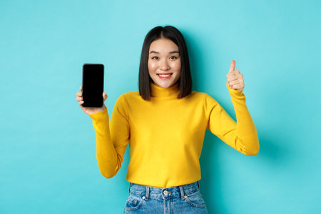 Technology and people concept. Cheerful asian girl in yellow sweater showing blank smartphone screen and thumbs up, demonstrate online offer, standing over blue backgroundの写真素材