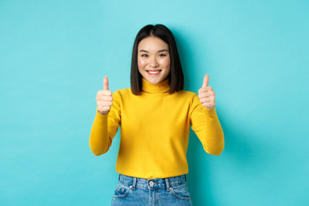 Cheerful asian female model showing thumbs up gesture, smiling and looking impressed, praise good product, standing over blue backgroundの写真素材
