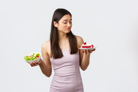 Healthy lifestyle, leisure and food concept. Beautiful asian girl staying on diet, pouting and looking with desire at piece cake, tempting to bite, holding salad another hand, white backgroundの写真素材