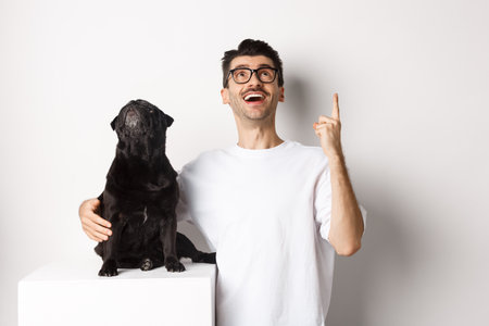 Image of happy young man and dog both looking up at promo, owner pointing finger at top, pug staring at loog, standing over white backgroundの写真素材