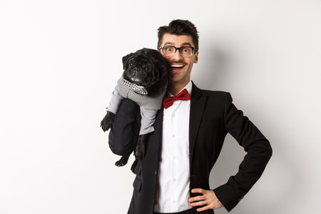 Handsome young man in suit and glasses holding cute black pug dog on shoulder, smiling happy at camera, wearing party outfits, white backgroundの写真素材