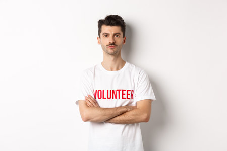 Serious young male volunteer in white t-shirt, holding arms crossed on chest, looking at camera, ready to helpの写真素材